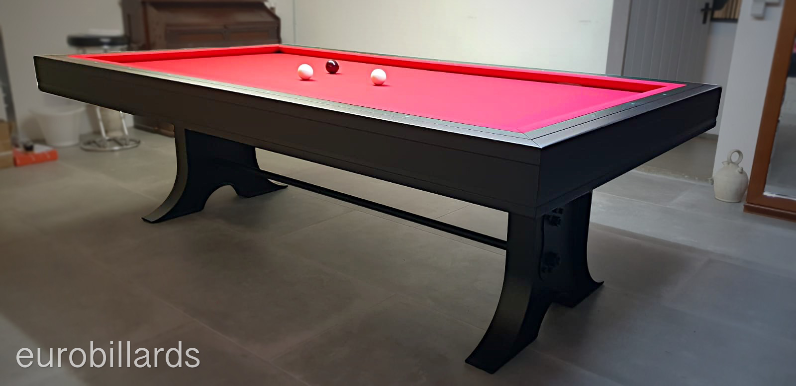 Le billard ATELIER finition noir mat monté en jeu français grace au système mixte XB qui remplace les bandes du jeu américain pose grave avec son tissu rouge soutenu par ses deux gros pieds en métal noir de style industriel.